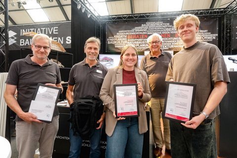 Kleine Gruppe mit Auszeichnungen posiert vor einer Ausstellungswand zum Thema Bauen und Planen.