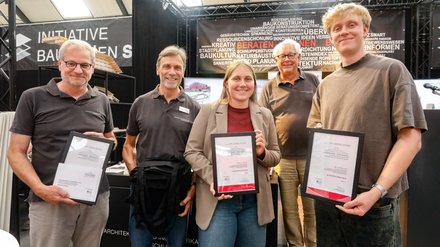 Kleine Gruppe mit Auszeichnungen posiert vor einer Ausstellungswand zum Thema Bauen und Planen.