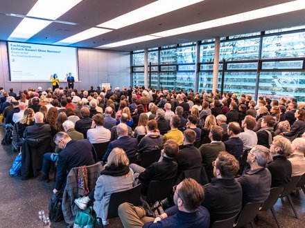 Zwei Personen stehen auf einer Bühne vor einer Leinwand mit Veranstaltungstitel; davor sitzt ein großes Publikum in Reihen, der Raum ist hell und modern gestaltet.