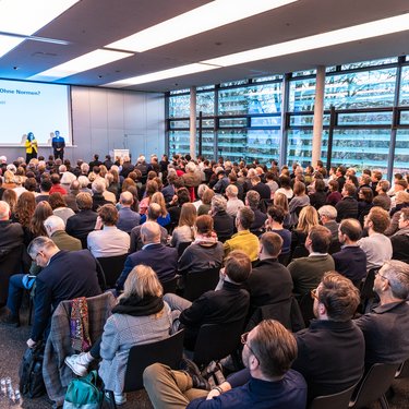Zwei Personen stehen auf einer Bühne vor einer Leinwand mit Veranstaltungstitel; davor sitzt ein großes Publikum in Reihen, der Raum ist hell und modern gestaltet.