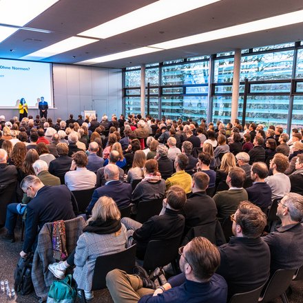 Zwei Personen stehen auf einer Bühne vor einer Leinwand mit Veranstaltungstitel; davor sitzt ein großes Publikum in Reihen, der Raum ist hell und modern gestaltet.