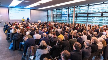 Zwei Personen stehen auf einer Bühne vor einer Leinwand mit Veranstaltungstitel; davor sitzt ein großes Publikum in Reihen, der Raum ist hell und modern gestaltet.