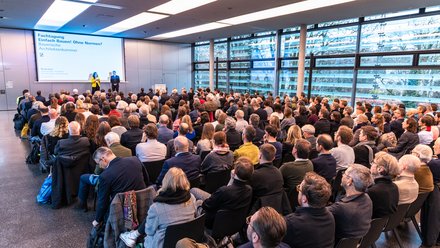 Zwei Personen stehen auf einer Bühne vor einer Leinwand mit Veranstaltungstitel; davor sitzt ein großes Publikum in Reihen, der Raum ist hell und modern gestaltet.