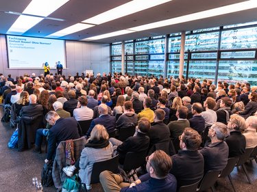 Zwei Personen stehen auf einer Bühne vor einer Leinwand mit Veranstaltungstitel; davor sitzt ein großes Publikum in Reihen, der Raum ist hell und modern gestaltet.