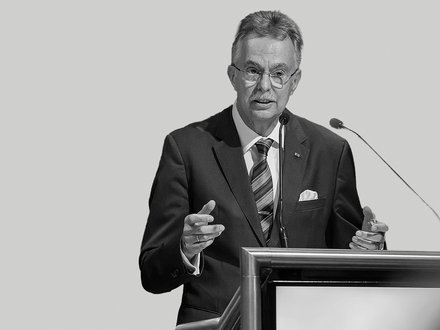 Schwarzweißfoto eines älteren Mannes in formeller Kleidung (Sakko, Krawatte, Einstecktuch) mit Brille und Schnurrbart, der gestikulierend an einem Rednerpult in ein Mikrofon spricht; auf dem Pult ist die Aufschrift „Architektenkammer Nordrhein‑Westfalen“ zu sehen.