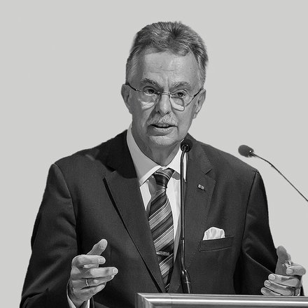 Schwarzweißfoto eines älteren Mannes in formeller Kleidung (Sakko, Krawatte, Einstecktuch) mit Brille und Schnurrbart, der gestikulierend an einem Rednerpult in ein Mikrofon spricht; auf dem Pult ist die Aufschrift „Architektenkammer Nordrhein‑Westfalen“ zu sehen.