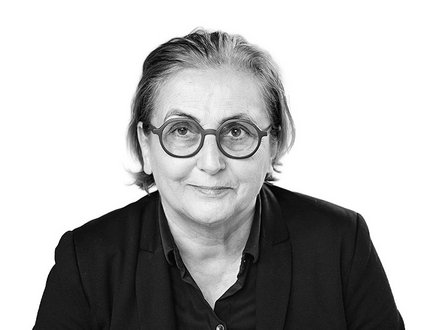 Schwarz-Weiß-Porträt einer Person mit schulterlangen, hellgrauen Haaren und runder Brille mit dunklem Rahmen. Sie trägt ein schwarzes Hemd und blickt direkt in die Kamera mit einem leichten Lächeln. Der Hintergrund ist weiß.