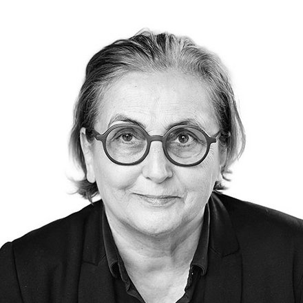 Schwarz-Weiß-Porträt einer Person mit schulterlangen, hellgrauen Haaren und runder Brille mit dunklem Rahmen. Sie trägt ein schwarzes Hemd und blickt direkt in die Kamera mit einem leichten Lächeln. Der Hintergrund ist weiß.