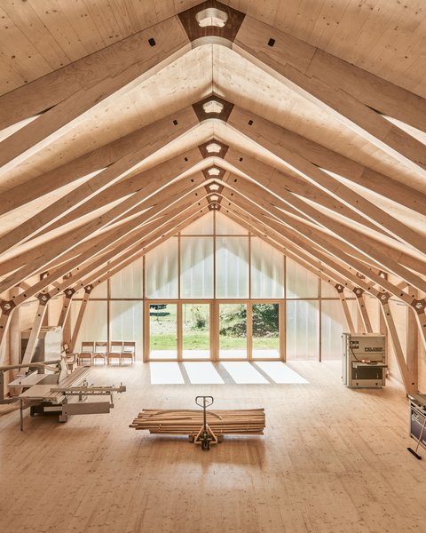 "Innenraum einer Holzhalle mit symmetrischer Dachkonstruktion aus hellem Holz. Das Dachgerüst zeigt Querbalken und Verbindungspunkte. Am Ende des Raums befindet sich ein großes Fenster mit Blick ins Grüne. Ein Foosball-Tisch und Stühle sind im Raum verteilt."