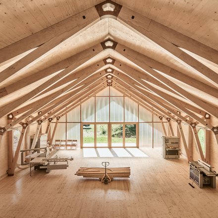 "Innenraum einer Holzhalle mit symmetrischer Dachkonstruktion aus hellem Holz. Das Dachgerüst zeigt Querbalken und Verbindungspunkte. Am Ende des Raums befindet sich ein großes Fenster mit Blick ins Grüne. Ein Foosball-Tisch und Stühle sind im Raum verteilt."