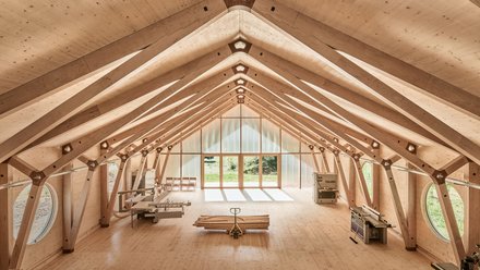 "Innenraum einer Holzhalle mit symmetrischer Dachkonstruktion aus hellem Holz. Das Dachgerüst zeigt Querbalken und Verbindungspunkte. Am Ende des Raums befindet sich ein großes Fenster mit Blick ins Grüne. Ein Foosball-Tisch und Stühle sind im Raum verteilt."