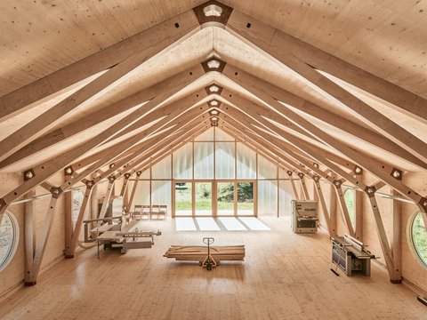 "Innenraum einer Holzhalle mit symmetrischer Dachkonstruktion aus hellem Holz. Das Dachgerüst zeigt Querbalken und Verbindungspunkte. Am Ende des Raums befindet sich ein großes Fenster mit Blick ins Grüne. Ein Foosball-Tisch und Stühle sind im Raum verteilt."