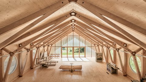 "Innenraum einer Holzhalle mit symmetrischer Dachkonstruktion aus hellem Holz. Das Dachgerüst zeigt Querbalken und Verbindungspunkte. Am Ende des Raums befindet sich ein großes Fenster mit Blick ins Grüne. Ein Foosball-Tisch und Stühle sind im Raum verteilt."