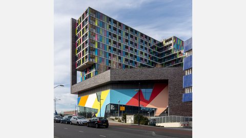 Perspektivische Straßenansicht eines modernen Gebäudekomplexes mit farbigen Fassadenelementen und großflächig gestalteter Sockelzone