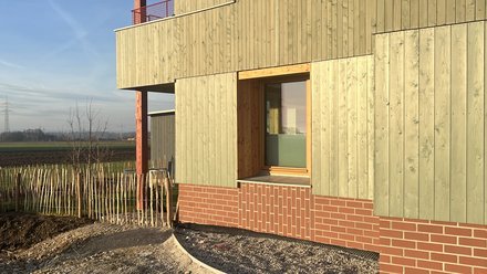 Mehrgeschossiges Wohngebäude mit vertikal strukturierter Holzfassade, tiefen Fensterlaibungen und Balkonen; Sockel aus Ziegelmauerwerk, im Vordergrund unfertige Außenanlagen.