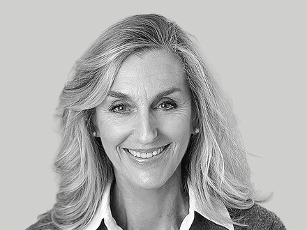 Schwarz-weißes Porträt einer erwachsenen Person mit schulterlangem, hellem Haar. Sie lächelt in die Kamera und trägt ein weißes Hemd unter einer grauen Strickjacke. Neutraler, hellgrauer Hintergrund.