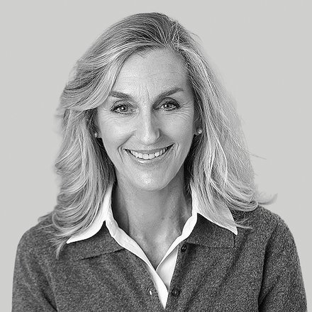 Schwarz-weißes Porträt einer erwachsenen Person mit schulterlangem, hellem Haar. Sie lächelt in die Kamera und trägt ein weißes Hemd unter einer grauen Strickjacke. Neutraler, hellgrauer Hintergrund.