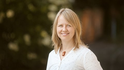 „Porträt einer lächelnden Frau mit schulterlangen blonden Haaren und weißer Bluse. Sie steht im Freien vor einem unscharfen, dunklen Hintergrund.“