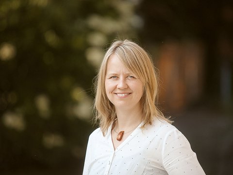 „Porträt einer lächelnden Frau mit schulterlangen blonden Haaren und weißer Bluse. Sie steht im Freien vor einem unscharfen, dunklen Hintergrund.“