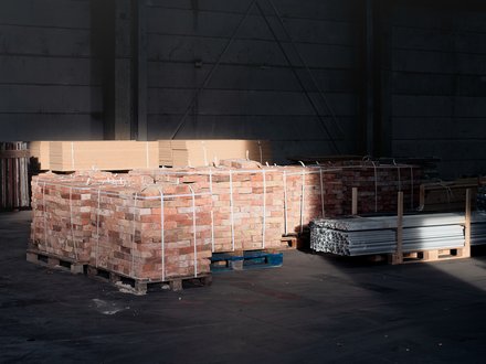 Paletten mit Ziegelsteinen und Metallprofilen in dunkler Lagerhalle, von einem Lichtstrahl teilweise beleuchtet.
