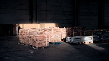 Paletten mit Ziegelsteinen und Metallprofilen in dunkler Lagerhalle, von einem Lichtstrahl teilweise beleuchtet.