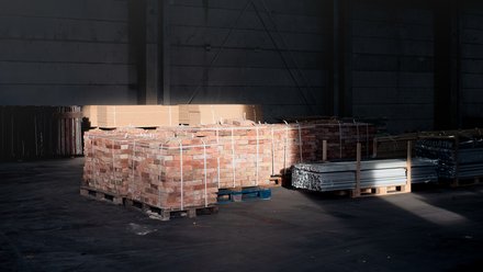 Paletten mit Ziegelsteinen und Metallprofilen in dunkler Lagerhalle, von einem Lichtstrahl teilweise beleuchtet.