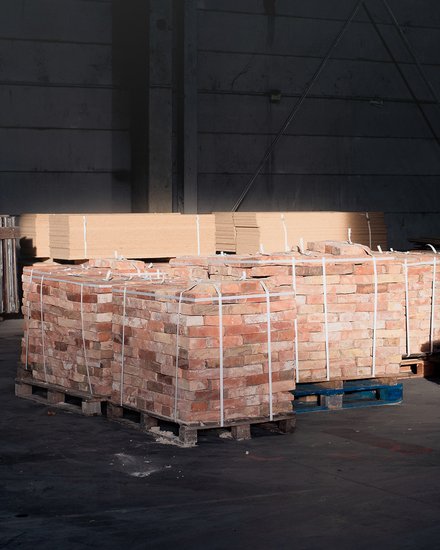 Paletten mit Ziegelsteinen und Metallprofilen in dunkler Lagerhalle, von einem Lichtstrahl teilweise beleuchtet.