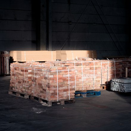 Paletten mit Ziegelsteinen und Metallprofilen in dunkler Lagerhalle, von einem Lichtstrahl teilweise beleuchtet.