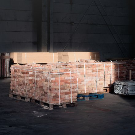 Paletten mit Ziegelsteinen und Metallprofilen in dunkler Lagerhalle, von einem Lichtstrahl teilweise beleuchtet.