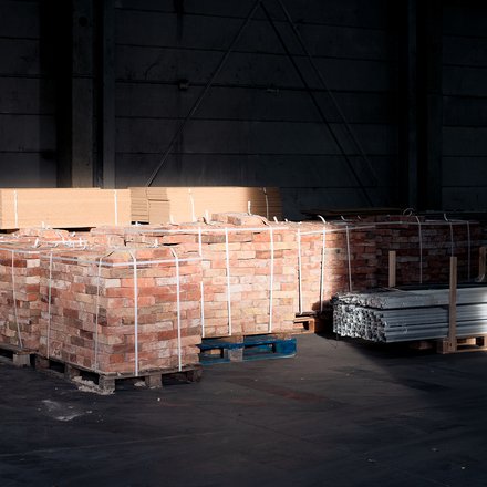 Paletten mit Ziegelsteinen und Metallprofilen in dunkler Lagerhalle, von einem Lichtstrahl teilweise beleuchtet.