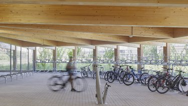 Innenansicht eines Holzparkhauses mit sichtbarer Holzbalkendecke, Fahrradabstellanlagen und einer Person in Bewegung im Vordergrund.