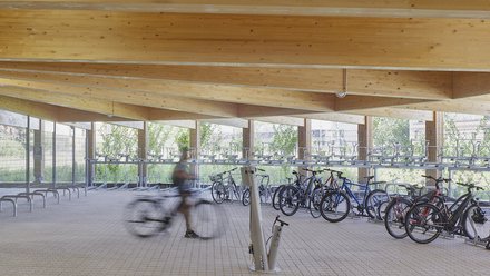 Innenansicht eines Holzparkhauses mit sichtbarer Holzbalkendecke, Fahrradabstellanlagen und einer Person in Bewegung im Vordergrund.