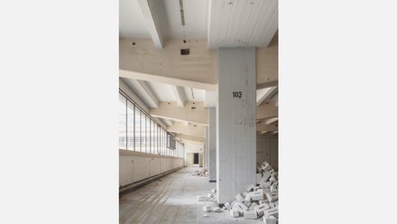 Innenraum während des Rückbaus  Ein langer Flur mit Betonstützen und Fensterband auf der linken Seite. Auf dem Boden liegen Schutt und herausgebrochene Mauersteine. Eine Stütze ist mit einer Nummer beschriftet. Das Bild dokumentiert den Rückbauprozess. Es zeigt, wie Teile des Bestands entfernt werden, um Platz für neue Nutzungen zu schaffen.