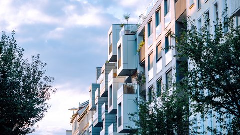 Moderne Wohnhäuser mit versetzt gestapelten, auskragenden Balkonen und viel Glas; Bäume säumen die Straße im warmen Abendlicht.