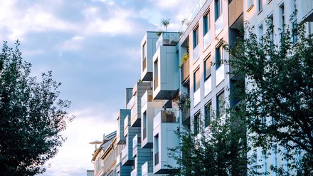 Moderne Wohnhäuser mit versetzt gestapelten, auskragenden Balkonen und viel Glas; Bäume säumen die Straße im warmen Abendlicht.