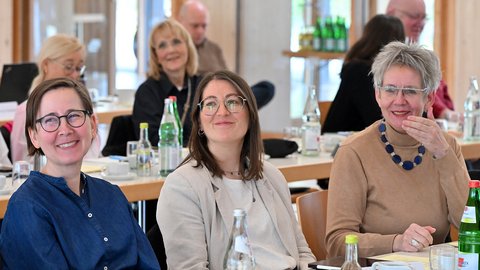 Drei Frauen mit Brillen sitzen an einem Tisch mit Wasserflaschen und Gläsern in einem hellen Raum