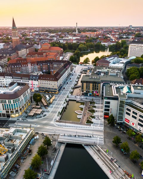 Luftaufnahme des neu gestalteten Holstenfleets in Kiel mit Wasserflächen, Fußgängerbereichen und umliegender Innenstadt; im Hintergrund ist die Kieler Stadtsilhouette zu sehen.