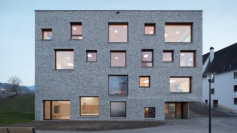Kubischer, mehrstöckiger Bau mit unregelmäßig angeordneten quadratischen Fenstern in einer hellgrauen Fassade vor bewaldeter Hügellandschaft