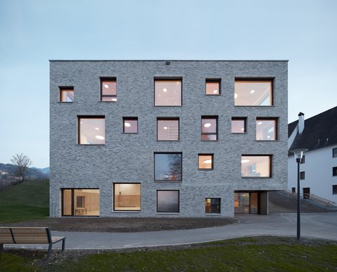 Kubischer, mehrstöckiger Bau mit unregelmäßig angeordneten quadratischen Fenstern in einer hellgrauen Fassade vor bewaldeter Hügellandschaft