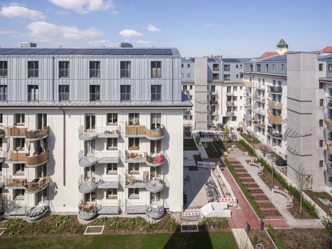 „Blick von oben auf eine moderne Wohnanlage mit hellem Putz, grauem Metalldach inklusive Photovoltaikanlage und Balkonen mit Holzverkleidung um einen begrünten Innenhof.“