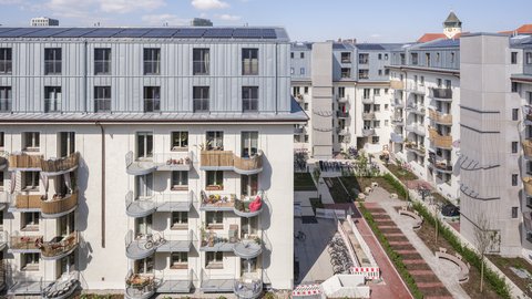 „Blick von oben auf eine moderne Wohnanlage mit hellem Putz, grauem Metalldach inklusive Photovoltaikanlage und Balkonen mit Holzverkleidung um einen begrünten Innenhof.“