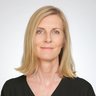 Porträt einer Person mit schulterlangem blondem Haar in schwarzer Bluse vor hellem, neutralem Hintergrund; ruhiger Blick in die Kamera.
