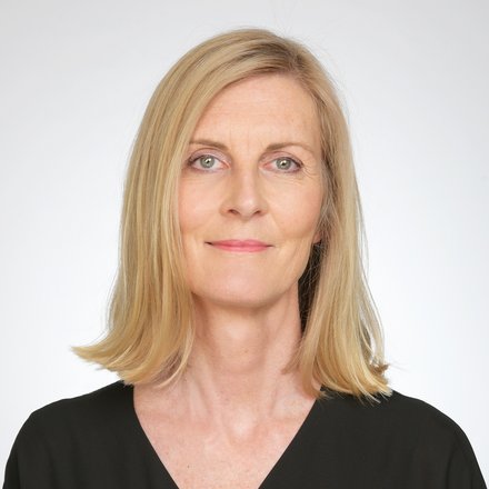 Porträt einer Person mit schulterlangem blondem Haar in schwarzer Bluse vor hellem, neutralem Hintergrund; ruhiger Blick in die Kamera.