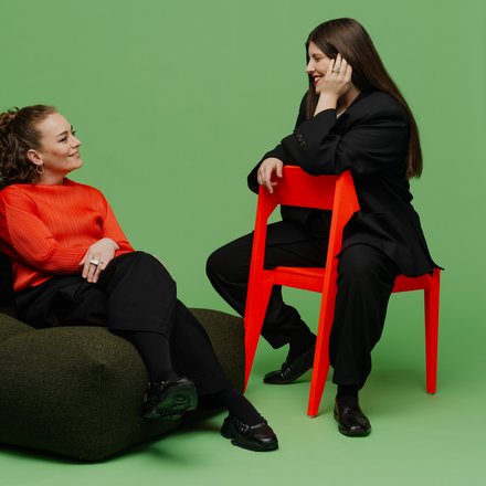  Porträt von zwei Frauen (Katharina Benjamin und Angelika Hinterbrandner) in einem Fotostudio mit grünem Hintergrund. Die linke Frau sitzt in einem leuchtend orangefarbenen Oberteil auf einem dunklen Sitzsack, die rechte Frau trägt einen schwarzen Oversize-Anzug und sitzt auf einem knallroten Stuhl. Beide lächeln sich an.