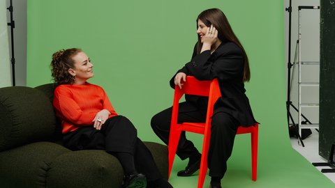  Porträt von zwei Frauen (Katharina Benjamin und Angelika Hinterbrandner) in einem Fotostudio mit grünem Hintergrund. Die linke Frau sitzt in einem leuchtend orangefarbenen Oberteil auf einem dunklen Sitzsack, die rechte Frau trägt einen schwarzen Oversize-Anzug und sitzt auf einem knallroten Stuhl. Beide lächeln sich an.