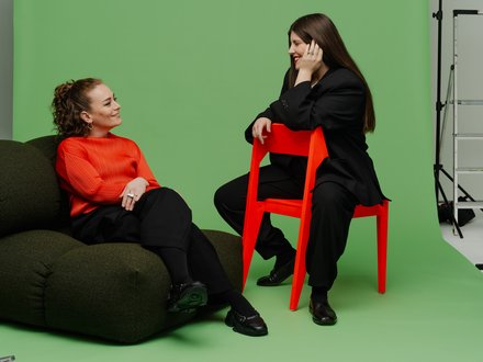  Porträt von zwei Frauen (Katharina Benjamin und Angelika Hinterbrandner) in einem Fotostudio mit grünem Hintergrund. Die linke Frau sitzt in einem leuchtend orangefarbenen Oberteil auf einem dunklen Sitzsack, die rechte Frau trägt einen schwarzen Oversize-Anzug und sitzt auf einem knallroten Stuhl. Beide lächeln sich an.