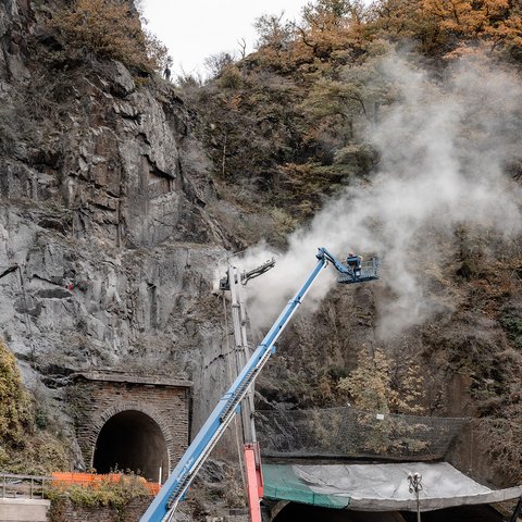 Felssicherungsarbeiten an einer steilen Felswand über alten Ziegelsteintunneln: Ein Arbeiter in einem Hubsteiger bohrt am Gestein und erzeugt eine große Staubwolke.