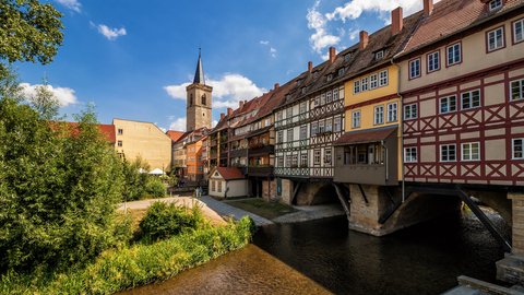 Eine historische Altstadtansicht zeigt dicht gereihte Fachwerkhäuser entlang eines Flusses, die Gebäude stehen teilweise auf Pfeilern im Wasser, ein Kirchturm ragt über die Dächer hinaus, wodurch das Bild die enge Verbindung von Stadt, Wasserlauf und Bauweise sichtbar macht und für gewachsene Stadtstrukturen und kulturelle Identität steht.