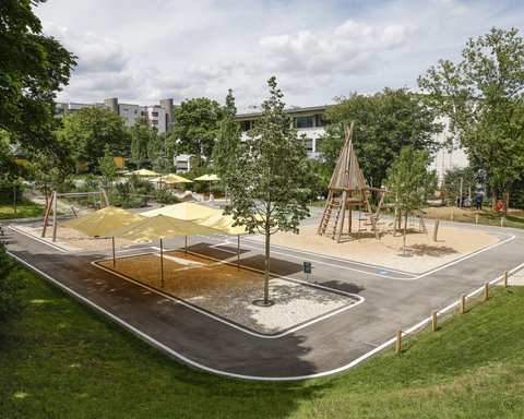 Spielplatz mit Sandfläche, Klettergerüst aus Holz in Form eines Tipis, mehreren Sonnensegeln und umgebenden Bäumen in einem Wohngebiet