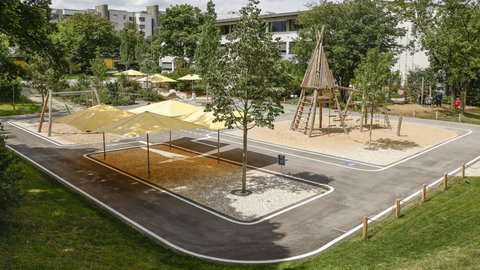 Spielplatz mit Sandfläche, Klettergerüst aus Holz in Form eines Tipis, mehreren Sonnensegeln und umgebenden Bäumen in einem Wohngebiet